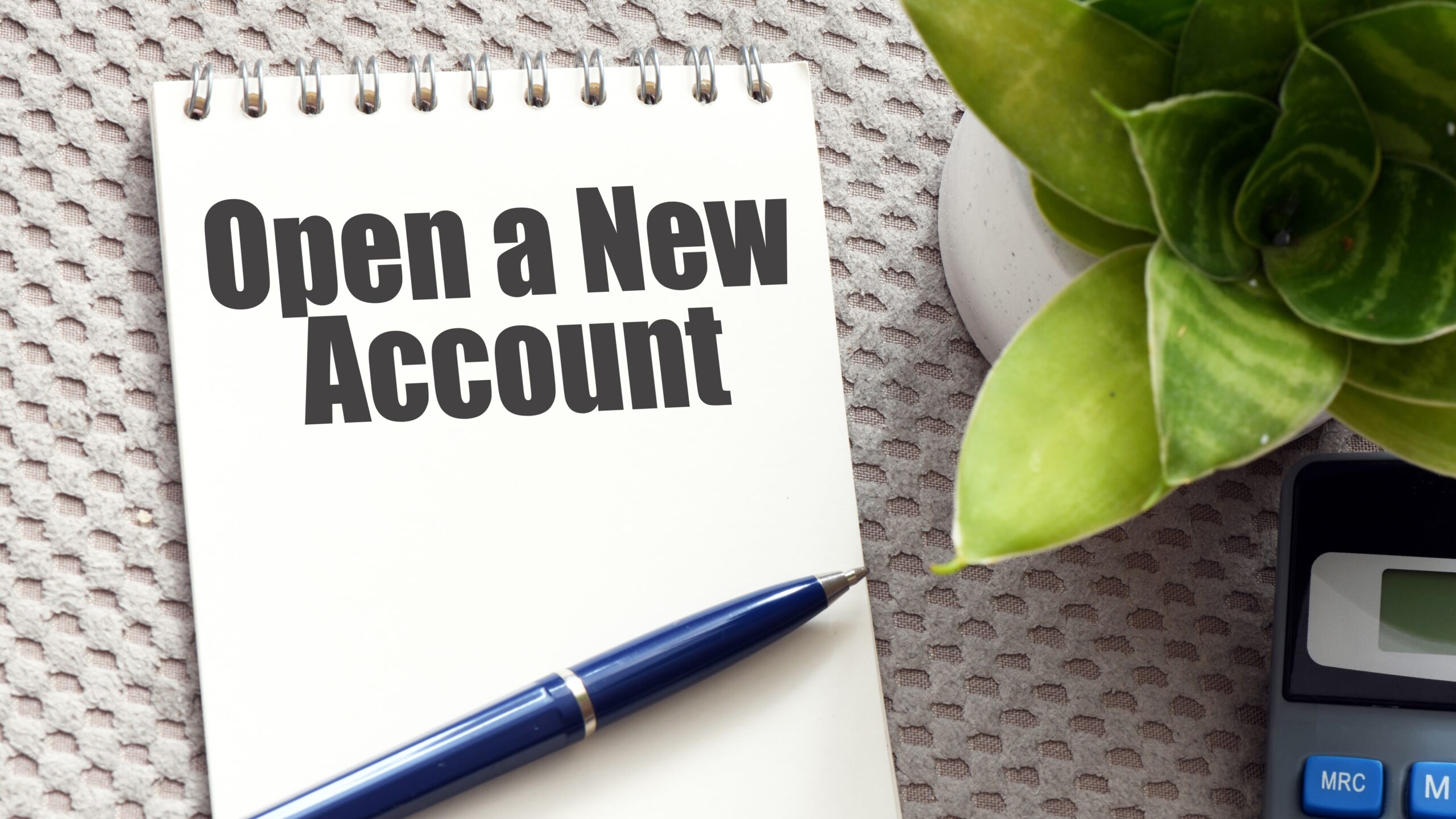 A spiral-bound notepad displays the bold, black text "Open A New Account." A blue pen rests diagonally across the bottom of the notepad, suggesting the action of writing or signing. The notepad sits on a textured gray surface, with a small potted succulent plant visible in the upper right corner and a portion of a calculator in the lower right. The overall composition implies a call to action for account creation.