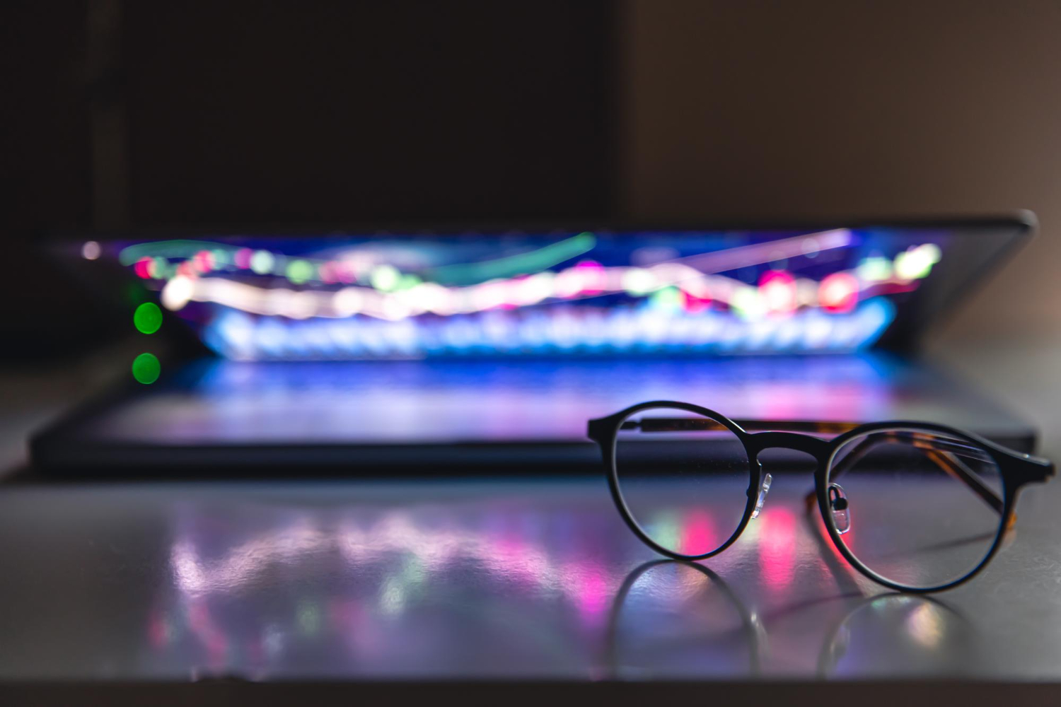 A pair of dark-rimmed eyeglasses rests on a reflective surface in front of a slightly open laptop displaying a colorful, glowing stock market or financial chart with dynamic lines and points of light.